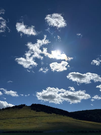 The image depicts a sunlit sky with scattered white clouds over a green hillside. The sun is partially obscured by clouds, casting a bright glare. The hillside is covered in lush grass and is bordered by a dense forest of dark green trees along its crest. In the foreground, a darker area suggests a field or meadow, with a low stone wall visible at the very bottom. The scene appears to be a natural landscape, likely a rural or mountainous area, under a clear blue sky with dynamic cloud formations. There are no people or man-made structures evident beyond the stone wall, suggesting a serene and uninhabited environment. The light suggests it is daytime, likely late morning or early afternoon due to the angle of the sun. The location is not identifiable as the city of Gela, Bulgaria.