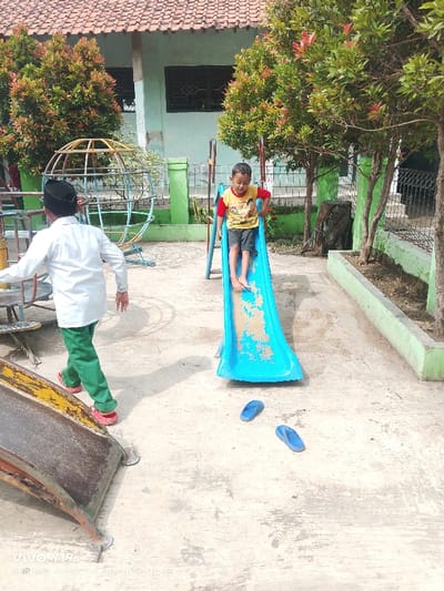 A child in a yellow and red shirt is sliding down a blue slide in a playground. A person in a white shirt and green pants walks to the left. Two blue flip-flops lay on the ground near the slide. The playground features other equipment, including a metal globe structure. Green bushes and trees are in the background, alongside a green building with a red roof. The scene appears to be outdoors, on a concrete surface.