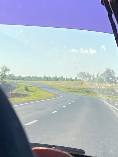The image shows a view from inside a vehicle looking out through a windshield onto a two-lane asphalt road that curves to the left. The road has white dashed lines marking the lanes. On either side of the road are grassy fields with scattered trees and bushes. In the distance, a line of trees forms a horizon. The sky is clear with a few wispy clouds. The upper portion of the windshield has a purple tint. The foreground of the image is dominated by a dark, blurry shape, likely the interior of the vehicle, with a red object visible at the bottom. The scene is outdoors, likely during daytime, given the bright lighting. The location is identified as Заглядовка, Russia.