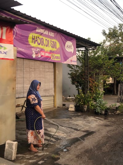 The image shows an outdoor scene at what appears to be a small business or shop in Sentul, Indonesia, during daylight hours, possibly late afternoon based on the lighting. A woman wearing a blue hijab and a patterned dress is standing on a wet, paved surface, holding a green hose and spraying water. She is barefoot. To her left, a building with a rolled-down bamboo-like shutter is visible. Above the shutter, a large, vibrant purple banner advertising "JARINGAN AXIS" is displayed, with yellow text that reads "HADIR DI SINI" and smaller text related to data packages and pricing ("3GB 30 HARI", "MULAI DARI 35 RIBU"). A portion of another banner, red and white with the word "AXIS" in blue, is visible to the far left. To the right of the building, there are various potted plants and trees, suggesting a somewhat green environment. Overhead, a tangle of electrical wires is prominent against the bright sky. The ground is wet and reflective, indicating recent watering or rain.