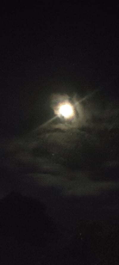 The image shows a night scene in Blangcot Tunong, Indonesia. A bright, partially obscured moon is the central focal point, surrounded by clouds. The foreground is dark and indistinct, possibly indicating land or trees.