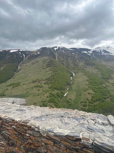 The image depicts a mountain landscape with a cloudy sky. In the foreground, there is a low, weathered stone wall constructed from irregularly shaped rocks of varying colors, including grey, orange, and rust tones, cemented together with a rough, light-colored mortar. To the left of the wall, a circular stone platform with a black metal railing is visible, overlooking a vast green mountain slope. The mountain itself is covered in lush green vegetation, with patches of trees and scattered snow on its upper reaches and along some gullies. The overall scene suggests an elevated viewpoint, likely from a historical structure or viewpoint in Gergeti, Georgia, offering a panoramic view of the surrounding mountainous terrain. No people are present in the image.