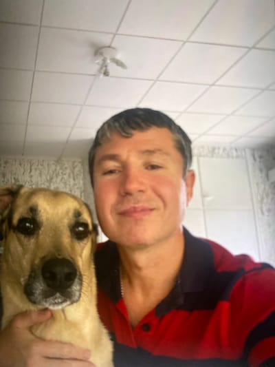 A man and a dog are in the frame, likely taking a selfie. The man has short, dark hair and is wearing a red and blue striped polo shirt. He is smiling. The dog, which appears to be a medium-sized breed with tan fur, is positioned in front of the man, looking towards the camera. Its head is partially obscuring the man's face. The background shows a white, tiled ceiling with a light fixture, and a wall with what appears to be patterned wallpaper. The scene suggests an indoor setting, possibly a home. There is no visible indication of the city of Kerava, Finland.