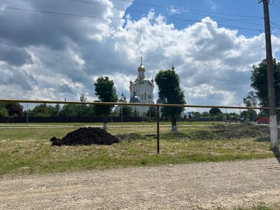 A white church with golden domes is visible behind a yellow horizontal pipe and some trees. The sky is cloudy with patches of blue. In the foreground, there is a pile of dark soil and patchy grass. A gravel road is in the bottom of the image. The church is located in the city of Dondukovskaya, Russia.