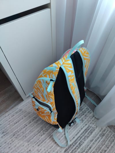 A backpack with an orange and blue palm leaf pattern rests on a light gray and white striped rug. The backpack is leaning against a white cabinet, and sheer white curtains hang in the background. The image appears to be taken indoors.