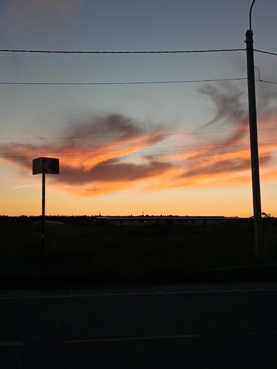 A twilight scene captures a rural landscape. The sky is a gradient of blues and oranges, with wispy clouds illuminated by the setting sun. A power line stretches horizontally across the upper portion of the frame. To the left, a traffic sign with a white chevron indicating a curve to the left is mounted on a pole with yellow and black stripes. In the mid-ground, a dark silhouette of trees and buildings is visible against the horizon. A dark road occupies the foreground, with a yellow line marking the lane. A concrete utility pole stands to the right of the frame. The image appears to be taken near Воскресенское, Russia, based on the provided context.