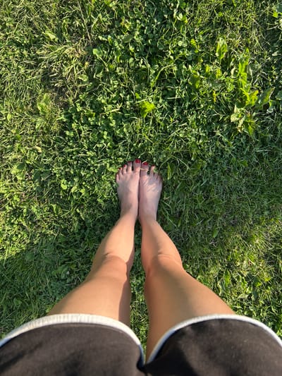 An overhead, full-body shot captures a person standing barefoot on a patch of green grass. The individual's legs, from mid-thigh down, are visible. They are wearing dark shorts with a white trim. Their feet are bare, with toenails painted red. The grass appears well-maintained, with a mix of clover and other green blades, illuminated by bright sunlight casting shadows. The scene is set outdoors, likely in a park or a backyard, in the village of Vyshevyichi, Ukraine.