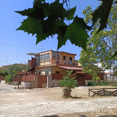 In Grumo Appula, Italy, a two-story house with a terracotta-tiled roof and yellow-painted exterior is visible, framed by lush green foliage. The house features a sunroom with horizontal blinds on the upper floor, accessible by an external staircase. A balcony wraps around the upper level, adorned with metal railings. Laundry hangs from a clothesline on the balcony, with a blue towel and a white garment visible. The property is enclosed by a stone wall topped with a brown metal fence. A small tree stands near the entrance. The ground in front of the house is a mix of dirt and stone paving. The sky is a clear, bright blue. A wooden fence is situated in the foreground. A timestamp in the bottom right corner reads "16.07.2026 14:13."