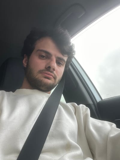 A man is in a car. The man has dark hair and a beard. He is wearing a white sweatshirt and a seatbelt. The interior of the car is dark. The sky is overcast. There is no indication of the location being Kvemo Rene, Georgia.