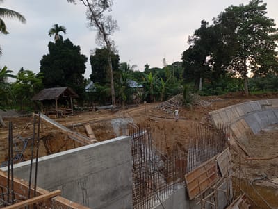 The image depicts a construction site in the city of Blangraleu, Indonesia, with a focus on civil engineering works. In the foreground, a substantial concrete wall is under construction, reinforced with a dense network of steel rebar. Wooden formwork is visible, indicating ongoing concrete pouring or preparation. The site is characterized by excavated earth and piles of gravel and rocks. A makeshift thatched-roof shelter is situated in the mid-ground, adjacent to a wooded area. Further into the background, several houses with dark roofs can be seen nestled amongst lush green vegetation, typical of a tropical environment. A lone individual, wearing a light-colored shirt and trousers, is standing on a higher level of the construction site, observing the work. The sky is overcast, suggesting a daytime scene. The construction appears to involve the creation of a retaining wall or a water channel, given the curved concrete structure visible in the upper right portion of the image. The overall impression is one of ongoing development and infrastructure work within a natural, tropical setting.