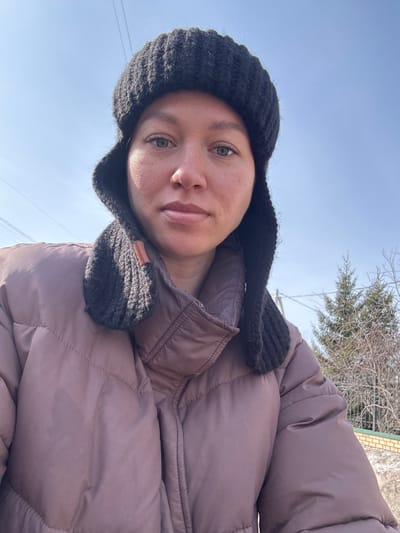 A close-up, eye-level outdoor portrait depicts an adult female with light-colored eyes and a neutral expression, positioned centrally. She is wearing a dark, possibly black, knit hat with ear flaps, one of which displays a small brown leather-like tag. Her torso is clad in a puffed, quilted jacket of a muted reddish-brown or mauve color. The background, consistent with a residential or suburban area in Gamovo, Russia, features a clear, bright blue sky in the upper quadrants. Several thin utility wires traverse the sky in the upper left. To the mid-right, a green coniferous tree is visible, flanked by bare deciduous trees and shrubs, indicating a non-summer season. In the lower right background, a dark metal fence or railing stands in front of a low brick wall with a lighter base and a darker top course. The scene is illuminated by natural, bright sunlight.