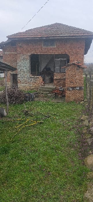 The image depicts the exterior of a two-story, brick-built structure located in Tsonevo, Bulgaria, under an overcast sky. The house features a reddish-brown tiled roof. The ground floor facade includes a large, recessed central archway that exposes a dark interior, suggesting an absence of a door or wall section. To the left of the archway, a rectangular doorway is visible. To the right of the arch, a protruding brick structure, also with a tiled cap, stands partially ruined. The brickwork across the entire structure shows signs of age, deterioration, and exposed mortar. A single rectangular window is present on the upper floor, positioned above the central arch.

The immediate foreground consists of green grass with interspersed yellowed vegetation, small purple flowers, and scattered branches or woody debris. To the left of the central arch, a pile of dark, thorny branches or shrubs is visible. Additional loose bricks and rubble are present at the base of the house, particularly near the central entrance. A metal fence post and wire fencing are partially visible on the far right, alongside a stone boundary. A thin, dark utility line spans diagonally across the upper portion of the grey, cloudy sky. No people or animals are present in the scene, and no specific actions or interactions are depicted.