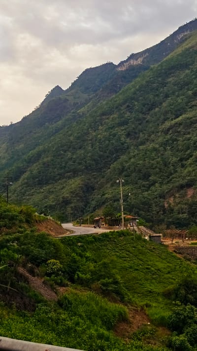 The image depicts a mountainous landscape in Minh Tân Commune, Vietnam, under an overcast sky. A winding road cuts through the lower portion of the frame, with a small building or rest stop situated on a curve. A utility pole stands next to the road. The mountainside is densely covered with trees. Grassy vegetation dominates the immediate foreground.