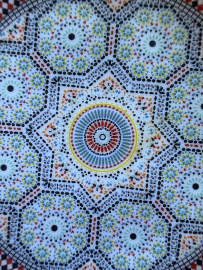The image displays a detailed, overhead close-up of a vibrant and intricate geometric pattern, characteristic of Moroccan decorative art. The central feature is a circular motif comprising concentric rings of small red, blue, and yellow dashes, encircled by a thin yellow line. Radiating outwards from this core are complex symmetrical designs formed by numerous small black dots, black curvilinear shapes resembling petals, and clusters of tiny colored elements in red, orange, yellow, light blue, and light green, set against a predominantly white or off-white background.
The pattern is highly repetitive and tessellated, featuring prominent multi-pointed star shapes formed by black dots and small light green star-like elements. These larger star patterns are interconnected by intricate lattices of black dots and dashes. The overall design suggests a traditional mosaic or painted ceramic surface, possibly a plate, tile, or a decorative panel, consistent with the location context of Loudaya, Morocco. There are no people, visible activities, or text present in the image. The lighting appears even, highlighting the bright, yet subtly textured, surface of the patterned object.