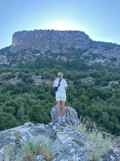 An adult stands on a rocky outcrop, facing away from the camera, looking towards a massive, steep cliff face. The cliff is dotted with numerous ancient rock-cut tombs, or necropolis, characteristic of the area. The cliff is covered in sparse vegetation, with a dense forest of green trees extending outwards from its base and filling the valley below. The sky is clear blue, with the sun's rays creating a bright halo behind the top of the cliff. The person is wearing a white t-shirt, white shorts, a white bandana, and a black sling bag. The person's hair is braided and falls down their back. The image was taken in the vicinity of Tlos Ancient City in the Seydikemer district of Turkey.
