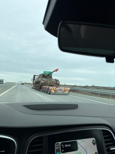 The scene is captured from the interior of a vehicle on the A1 highway (Trakia motorway) in Bulgaria, near the exit for the city of Svoboda, as indicated by a green road sign displaying "Св. Свобода" (Svoboda) and "Бургас" (Burgas). The current time displayed on the vehicle's infotainment screen is 12:02, showing a remaining distance of 155 km on the A1 highway, with a partial speed limit indicator of 105 km/h and a 5G network signal.
In the midground, a multi-axle heavy transport truck is observed in an adjacent lane, travelling in the same direction. The truck's flatbed trailer carries a large, camouflaged self-propelled howitzer. The howitzer's main gun barrel is angled upwards and forward, fitted with an orange protective cap. The rear of the truck's trailer displays red and white diagonal reflective safety markings and a circular yellow sign indicating a 30 km/h speed limit for oversized loads.
The highway features multiple lanes with white line markings. A metal guardrail separates the roadway from the undeveloped land on the right side. The background consists of a flat, open landscape under an overcast sky, with sparse vegetation visible along the horizon. No individuals are explicitly visible in the frame, though the perspective implies a driver or passenger taking the photograph.
