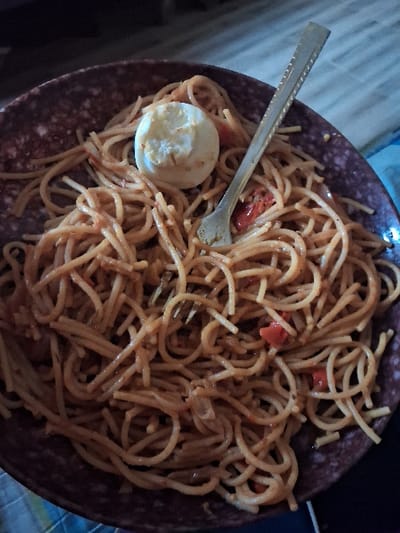 The image displays a meal of spaghetti served in a dark brown bowl with a speckled pattern. The spaghetti is coated in a reddish-orange sauce, visibly containing small red fragments (likely tomatoes) and translucent white-yellow pieces (possibly onions). A single, peeled, hard-boiled egg, exhibiting a slightly irregular surface, is centrally placed on top of the spaghetti. A silver-colored fork, featuring a textured design on its handle, is inserted diagonally into the pasta. The bowl rests on a light-colored, possibly patterned surface, with the background consisting of indistinct, darker, out-of-focus shapes. The scene is illuminated by dim light. This depiction is located in Ukwulu, Nigeria.
