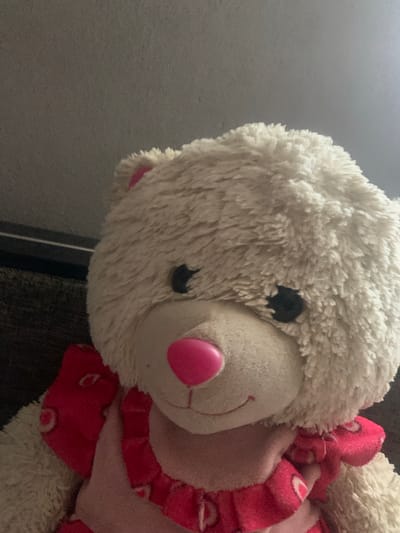 This is a close-up, downward-angled, eye-level, medium shot of a fuzzy white stuffed animal, possibly a bear or sheep, with a pink nose and black button eyes. The stuffed animal is wearing a bright pink, ruffled accessory that appears to be a collar or scarf decorated with a repeating pattern of white hearts. The background is a dark, textured surface, likely a piece of furniture. The lighting is soft and diffused, suggesting an indoor setting. There are no people or text visible in the image. The focus is entirely on the stuffed animal and its details.