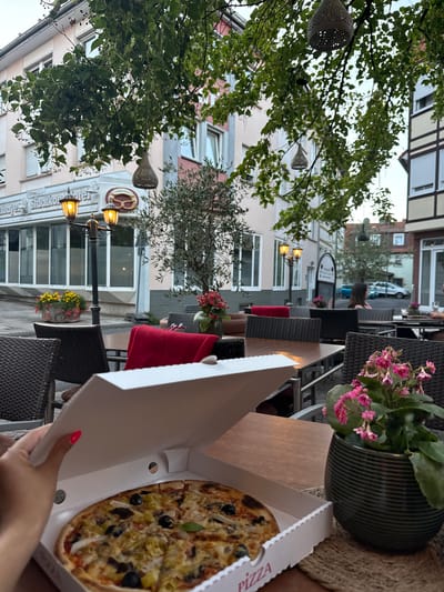 The image depicts an outdoor dining area of a restaurant in Bad Orb, Germany, likely in the evening. A hand with bright pink nail polish is opening a pizza box, revealing a whole pizza with olives and what appears to be artichoke. In the foreground, there is a wicker chair and a wooden table. Behind the pizza, more tables and chairs are visible, some occupied by patrons. A potted plant with pink flowers sits on a table. The background shows buildings, some with half-timbered elements, suggesting a traditional German town setting. Lampposts and decorative hanging lanterns are also present. The overall atmosphere is relaxed and inviting, indicative of a casual dining experience.