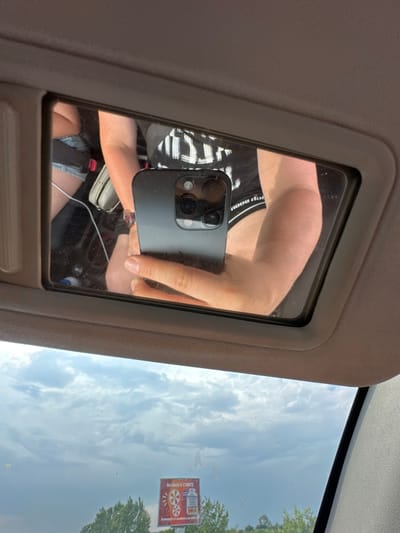 The image shows a person taking a selfie in a car's vanity mirror. The person is holding a black smartphone with a visible camera lens array. They are wearing a black and white striped shirt and denim shorts. The reflection in the mirror also shows a seatbelt. Outside the car, through the windshield, the sky is visible with scattered clouds. A small sign with some text and a graphic of gears is affixed to the windshield, likely a sticker or toll pass. The time of day appears to be daytime, with bright lighting suggesting fair weather. The setting is inside a car, likely on a road trip given the visible scenery outside. There is no clear indication of the person's emotions, but the act of taking a selfie can imply a casual or self-aware moment. The visible text on the sign is partially obscured and difficult to read, but some letters and numbers are discernible. The location context provided is Saraia, Bulgaria.