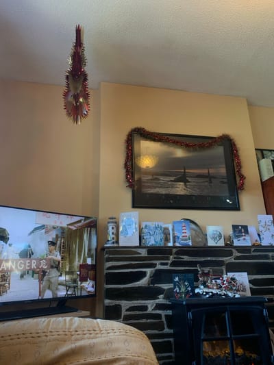 This image captures a domestic interior in Tideford, United Kingdom, depicting a living room with festive decorations.
A white, textured ceiling hangs over light beige walls. A red and metallic silver, conical spiral decoration hangs from the ceiling in the upper left quadrant.
Below it, a flat-screen television displays a scene featuring Asian individuals in what appears to be an outdoor market or street. One individual, possibly a guard or soldier, wears a light-colored uniform and a hat, holding a long object resembling a rifle. English text "ANGER" and accompanying Chinese characters, including "危," are visible on the lower-left of the screen. The bottom portion of a light-colored, textured upholstered furniture item, possibly an armrest or cushion, is present in the foreground, partially obscuring the base of the television and fireplace.
On the right side of the image, a fireplace structure constructed from dark, stacked stone occupies the center-right. A black-framed artwork, depicting a dramatic ocean scene with whales under a stormy sky, hangs above the mantelpiece. A red tinsel garland with small red spherical ornaments is draped over the top and sides of this artwork.
The stone mantelpiece holds a white and red lighthouse figurine on the left, followed by several upright greeting cards, predominantly Christmas-themed, featuring winter landscapes, a lighthouse, and other designs.
Below the mantelpiece, a black electric fireplace insert displays artificial flames behind a glass panel. On the top surface of this insert, a small card depicting snowdrops is placed next to a Christmas figurine of Santa Claus in a sleigh pulled by reindeer, laden with miniature gifts. Another small card is visible to the right of the figurine.