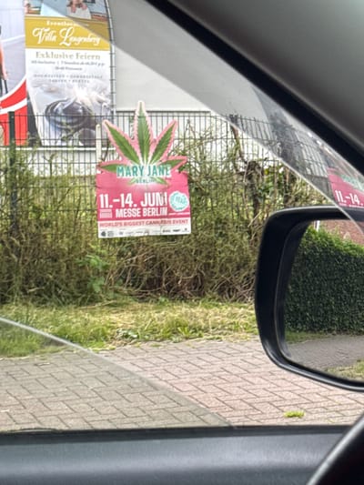 The image shows a promotional poster for "Mary Jane Berlin," described as the "World's Biggest Cannabis Event," which took place from June 11-14 in Berlin. The poster features a stylized cannabis leaf with pink and green colors and is attached to a fence. In the background, another poster with German text, likely advertising an event or venue, is visible. The scene appears to be outdoors, with grass and foliage surrounding the fence. The perspective suggests the photo was taken from inside a vehicle, with a car mirror visible on the right side of the frame. The presence of the "Mary Jane Berlin" poster strongly suggests the image was taken in Berlin, Germany, as indicated by the poster itself.