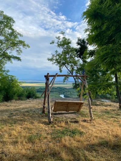 A handmade wooden swing set, constructed from rough-hewn logs and planks, is positioned on a dry, grassy hilltop overlooking a wide river valley. The swing seat is empty. Trees with lush green foliage frame the scene on either side. In the distance, the valley unfolds with patchwork fields and a meandering river, under a sky dotted with white clouds. The overall impression is one of rustic tranquility in a rural landscape, likely near Horești, Moldova.