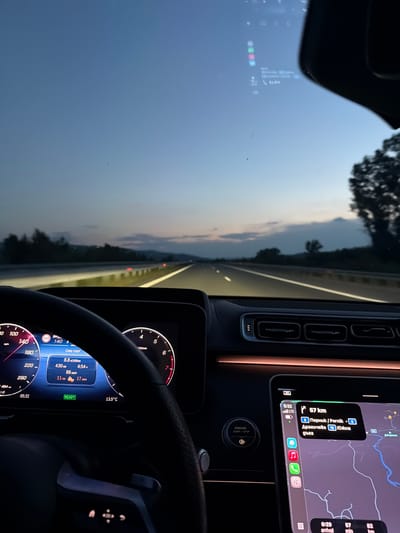 The image shows the interior of a car, focusing on the dashboard and windshield view. The car is in motion on a multi-lane highway at twilight. The speedometer reads about 140 km/h. The infotainment system displays data regarding the trip, showing 430 kilometers driven, with a fuel consumption of 8.8 liters per 100 km. The outside temperature is 13.5 degrees Celsius. A smartphone mounted near the dashboard is displaying a navigation app, showing the destination as Pernik, Bulgaria, 57 km away, accessible via road 6. Given the destination and the "Южна дъга" mentioned on the screen, it is unlikely the location is Mursalevo.