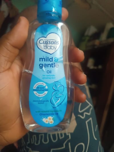 The scene depicts a clear plastic bottle of "Cussons Baby mild & gentle oil," approximately 3/4 full, held by a person's left hand. The hand, with brown skin, grips the bottle from the sides, with fingers visible on the left and right. The bottle features a light blue label displaying the brand name "Cussons Baby" within a heart shape, and text indicating "mild & gentle oil for naturally glowing skin." Further text details its use for "baby's skin" (all over moisturisation), "baby's hair," and states it is "Dermatologically tested," containing "natural ingredients - Chamomile Oil" with an illustration of chamomile flowers. The bottle has a light blue flip-top cap. The background is out of focus. To the lower right, a dark fabric with a lighter, possibly geometric, pattern is visible. The upper right background shows indistinct shapes and colors, suggestive of an indoor environment. This event occurs in Okere-Urhobo, Nigeria.