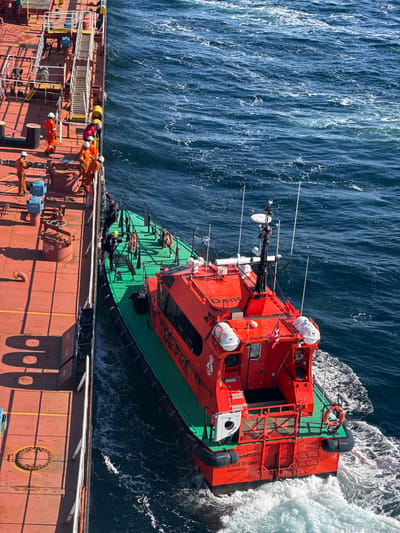 The image shows a pilot boat, the "DanPilot Bravo," alongside a large vessel at sea. Several individuals in orange jumpsuits are on the deck of the larger vessel, appearing to be crew members. Two individuals, one in a dark jacket and another in a yellow helmet, are in the process of climbing from the pilot boat onto the larger vessel. The pilot boat is bright orange with a green hull and has various antennas and equipment on its roof. The sea is choppy and blue, with white wakes indicating movement. The larger vessel has a red deck and metal railings. The scene appears to be a pilot transfer operation, a common maritime activity. The presence of "Svendborg" on the pilot boat suggests the location is near Svendborg, Denmark.