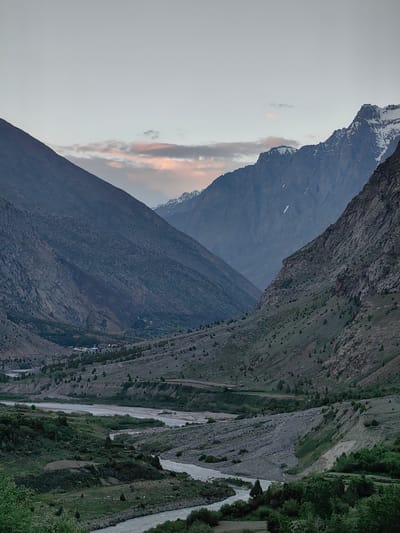 This media file depicts a serene mountain valley landscape at dusk. The scene is dominated by towering, rugged mountains on either side, with the left mountain slope featuring a rough, textured surface dotted with small, dark shrubs. The right side showcases a steeper, rockier incline. In the distance, snow-capped peaks rise, hinting at higher altitudes.
The valley floor is characterized by a winding river, its waters appearing calm and reflecting the muted light. Lush green vegetation covers the lower slopes and the riverbanks, interspersed with patches of rocky terrain. A small cluster of buildings, indicative of a village, is visible nestled within the valley, illuminated by faint lights. Roads or tracks are discernible winding through the landscape.
The sky is a soft, overcast grey, with a few wisps of pinkish clouds suggesting the lingering glow of sunset. The overall mood is tranquil and majestic, emphasizing the vastness and natural beauty of the mountainous region. The time of day is clearly evening, transitioning into night. There is no visible text in the media file. The location is Kead, India.