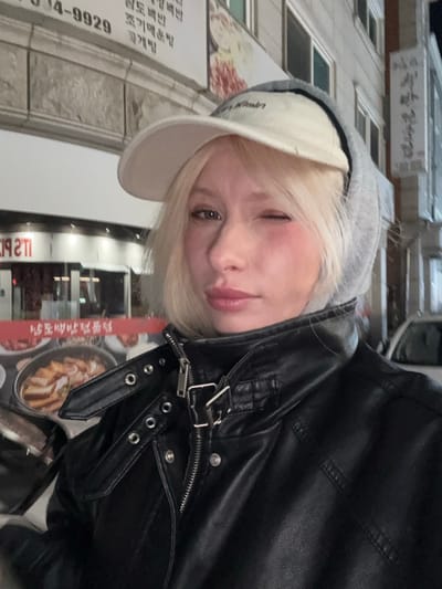 A blonde individual, appearing female, is centered in a medium-close-up selfie, captured at night in an urban setting identified as Namyangju-si, South Korea. The subject wears a light-colored baseball cap over a gray hooded sweatshirt, and a black faux leather motorcycle-style jacket with a zippered front and a buckled, studded collar strap. The individual's left eye (viewer's right) is closed in a wink, while the right eye is open, and the lips are slightly parted. The background is an illuminated street scene featuring multiple commercial establishments. To the immediate left and behind the subject, a storefront displays a red sign with partial English text "IT'S P..." and an accompanying sign showcasing images of Korean food dishes, including a large stew or braised meat platter. Above this, a white sign with Korean text includes a phone number (010-944-9929) and phrases related to real estate or business services. Further right, a vertical sign on a building facade displays the Korean characters "종가집." Artificial street and storefront lighting casts an ambient glow over the scene.