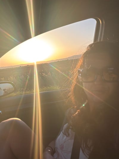 The image shows a woman sitting in a car with the sun setting in the background. She is wearing sunglasses and her hair is dark and long. The sun is low on the horizon and casting a warm glow over the landscape, which appears to be rural with fields and distant hills. The interior of the car is visible, including a side mirror reflecting another vehicle. The scene conveys a sense of a journey or a moment of reflection during a road trip. The location is stated to be in Zagortsi, Bulgaria.