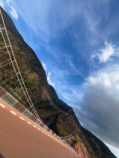 The image shows a low-angle view of a bridge in Tabaroucht, Morocco, with a mountainside and sky as a backdrop. The bridge is primarily reddish-brown, with white and red safety railings. White support cables extend from the bridge. A steep, rocky mountainside rises in the background, covered with sparse green vegetation. A mix of blue sky and clouds dominates the upper portion of the frame, with a section of darker, overcast clouds on the right.