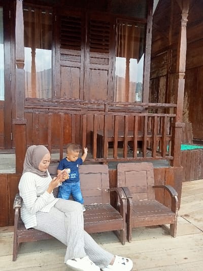 The image captures an outdoor scene on a wooden porch or balcony of a traditional brown wooden building in Kajar, Indonesia. In the foreground, an adult female is seated on a dark brown wooden chair to the left, dressed in a light brown hijab, a horizontally striped light-colored top, and light grey wide-leg pants, with white clogs on her feet. She is looking down, engaged with a pink-cased smartphone held in both hands. To her right, a toddler boy, wearing a dark blue t-shirt and blue denim shorts, stands barefoot, facing away from the camera. His left hand rests on a dark brown wooden railing that extends across the middle ground. Behind the railing, the building's facade is visible, featuring several window or door frames with dark brown wooden panels, some appearing to be louvered shutters, others containing glass panes that reflect the sky and show light-colored curtains within. An additional empty dark brown wooden chair is positioned to the right of the seated woman. The floor is composed of wide wooden planks.