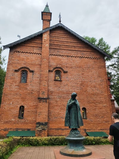In Selivanovo, Russia, a statue stands before a brick building. The statue depicts a person in a long cloak, holding their hands together in front of their chest. The building is constructed of red brick with arched windows and decorative brickwork along the roofline. A green metal roof covers a small section at the base of the building. Trees are visible behind and to the right of the building. A person wearing a black t-shirt and backpack stands to the right of the frame, facing away from the camera and towards the statue. The ground in front of the statue is paved with brick. The sky is overcast.