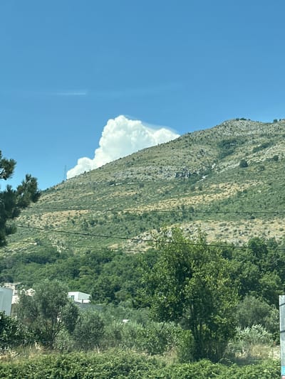 This is a vertical, medium shot photograph of a sunlit, mountainous landscape in Gornji Katun, Montenegro. The scene is dominated by a steep, rocky mountainside covered in sparse green vegetation. A large, fluffy white cloud is visible against a clear blue sky to the left of the mountain. A dense forest of green trees occupies the lower right portion of the image, obscuring much of the ground. A modern, white building with a flat roof is partially visible within the trees, suggesting a residential or possibly commercial structure. Power lines run diagonally across the image, hinting at the presence of infrastructure. The bright sunlight indicates it is daytime, and the clear sky suggests fair weather. The overall impression is one of natural beauty and rural habitation. No people are visible, and no text is present in the image.