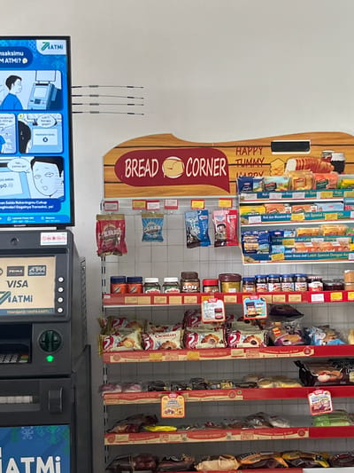 This image captures a close-up, interior view of a convenience store, likely a minimarket, in Meteseh, Indonesia. The scene is dominated by a wall-mounted display shelf filled with various packaged food items, primarily bread, pastries, and jars of spreads, arranged in neat rows. A digital display screen, likely for advertising or information, is positioned to the left of the shelves, showing a comic strip related to ATM transactions. Below the screen is an ATM machine, branded with "PRIMA" and "ATM Bersama." The overall lighting is bright and artificial, typical of an indoor retail environment. The focus is on the products and the transaction interfaces, suggesting a commercial setting.