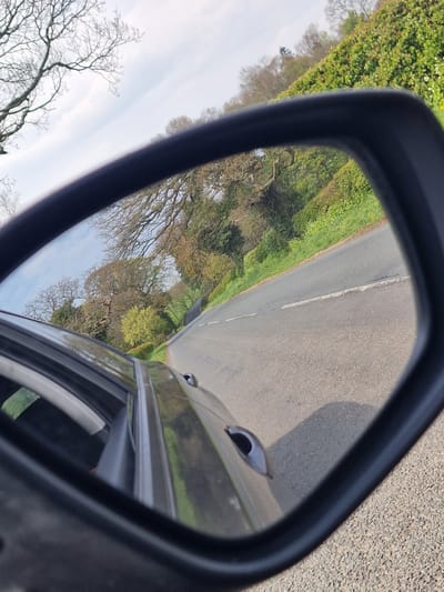 The image shows a view in a car's side mirror, reflecting a road alongside a grassy area, with trees in the background. The road is paved and marked with a single white line. Part of the car door and handle are visible in the lower right corner of the mirror. The sky is visible through the trees on the left. The photo appears to be taken in Framwellgate Moor, United Kingdom, as the surroundings are representative of the environment.