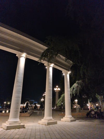 The image depicts an outdoor scene at night, featuring a colonnade with three prominent white pillars illuminated from above. The ground in the foreground is paved with interlocking grey bricks. Beyond the pillars, a park-like setting is visible, with trees, lampposts, and a body of water that reflects the lights. In the middle ground, there are several figures, some seated and others standing, suggesting a public gathering or recreational activity. In the background, a building with illuminated windows is discernible, and more lampposts are scattered throughout the area. The overall ambiance is that of a well-lit public space at night, possibly a garden or promenade in Ganja City, Azerbaijan.