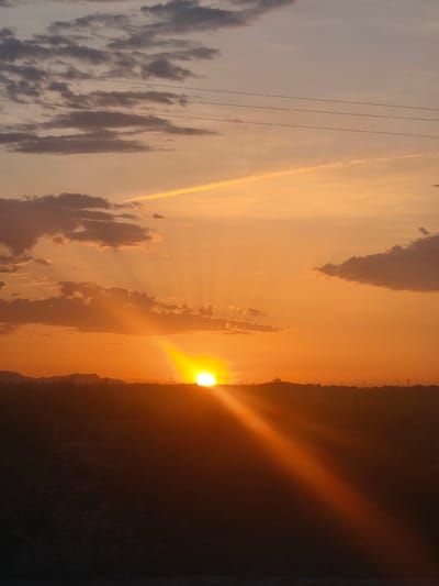 The image depicts a sunset over a rural landscape, likely on the outskirts of Riba-roja de Túria, Spain. The sun, a bright, intense orb, is setting near the horizon, casting long, golden rays across the sky. The sky is a gradient of warm oranges and yellows, transitioning to a softer peach and then a pale lavender towards the top. Wispy clouds, tinged with pink and gray, are scattered across the sky, with some larger, darker clouds present on the left side.

In the foreground, a dark, silhouetted landscape stretches across the bottom of the frame. This appears to be a sparsely vegetated area, possibly with some rocky terrain. The silhouette suggests rolling hills or a slight elevation. In the middle ground, a faint line of trees and structures can be discerned along the horizon, with what appear to be power lines stretching across the upper portion of the sky. There are no people or animals visible in the image. The overall impression is one of a tranquil, natural scene at the end of the day.