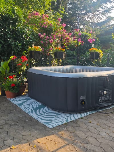 This media file captures an outdoor scene in what appears to be a garden setting, likely in Lügde, Germany. The primary object is a large, inflatable hot tub, dark grey with a light grey inflatable rim, resting on a patterned outdoor mat. The mat has a light blue and white leaf pattern and is placed on a paved patio made of interlocking grey stones.
To the left of the hot tub, there are several potted plants. One large plant with broad green leaves stands next to two smaller pots filled with red flowers and green foliage, likely geraniums. Behind these plants, a dense green ivy-covered wall forms the backdrop.
The background is dominated by lush greenery and flowering plants. A fence with decorative metalwork runs horizontally behind the potted plants, supporting hanging baskets filled with vibrant pink and orange flowers. Trees with sun-dappled foliage are visible in the upper right portion of the image, suggesting it is a sunny day. The warm, golden light suggests it is either early morning or late afternoon. The overall atmosphere is serene and inviting, indicative of a tranquil garden setting.
On the side of the hot tub, some text is visible, including "DO NOT OVER INFLAT" and instructions like "STEP 1," "STEP 2," "STEP 3," and "STEP 4," implying it is a portable hot tub with setup instructions. There are also control buttons and a dial on the unit. No people are present in the image.