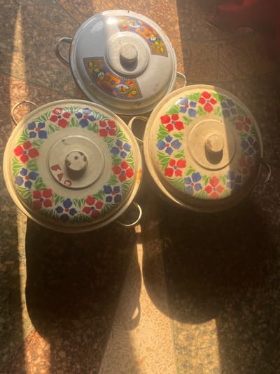 Three lidded, metallic serving pots are positioned on a tiled floor in Awgbu, Nigeria. The pots are round with side handles on the base and a central knob on each lid. The uppermost pot features a lid with a colorful, abstract pattern. The two lower pots have lids decorated with a repetitive floral motif, characterized by red and blue flowers interspersed with green leaves on a white background. The tiled floor consists of light-colored, possibly beige or cream, rectangular tiles with a speckled texture. Strong, directional sunlight illuminates the scene from the left, casting prominent, dark shadows of the pots onto the floor, extending towards the right. No individuals are present, and no actions or interactions are depicted.