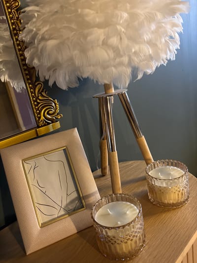 An interior scene in a room within the city of Баня, Bulgaria, depicts a collection of decorative objects arranged on a light-colored wooden surface against a solid dark teal wall. A large, unlit table lamp with a white feather shade stands on a tripod base composed of polished silver-colored metal and light wood sections. To the left, a portion of an ornate, antique gold-framed mirror is visible, tilted slightly, reflecting part of the feather lamp and a purple-hued area. In front of the mirror, a rectangular photo frame with a textured pale pink or beige border and a gold inner frame displays minimalist line art of a human figure. Two identical, unlit candles in amber-tinted, diamond-patterned glass holders are positioned on the wooden surface in front of the lamp's base.