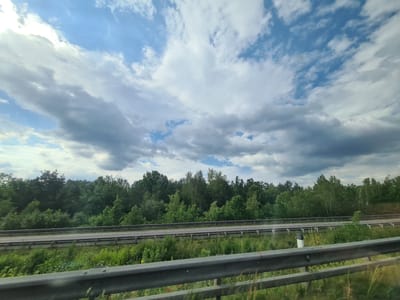 The image depicts a view of a highway bordered by a dense green forest under a cloudy sky. The highway has two lanes in each direction, separated by a metal guardrail. The foreground shows another guardrail, suggesting the photo was taken from a vehicle. The forest is lush and green, with a variety of trees. The sky is filled with a mix of white and grey clouds, with some patches of blue sky visible. The scene appears to be a daytime shot, possibly taken in the late spring or summer. There are no people or animals visible in the image. The location is identified as Loipersbach, Austria.