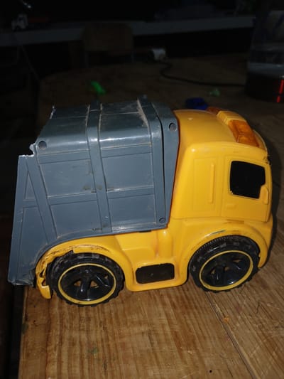 This is a media file of a yellow toy truck with a grey cargo bed. The truck is lying on its side on a wooden surface, with its wheels pointing upwards. The truck appears to be made of plastic and shows some signs of wear and tear, with a crack visible on the front fender. The background is dark and out of focus, but it appears to be an indoor setting, possibly a workshop or storage area. There are some blurry objects in the background, including what looks like a black electronic device and a small green object. The lighting is dim. The overall impression is a close-up shot of a used toy, likely in a home environment.