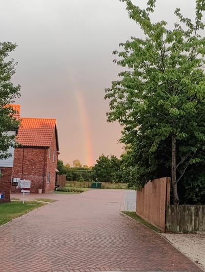 The image captures a street scene in the city of Mid Suffolk, United Kingdom, during what appears to be either sunrise or sunset, indicated by the warm, orange glow on the brick building and the soft, diffused light in the sky. A prominent rainbow is visible in the hazy sky, arcing from the distant horizon upwards.
In the foreground, a red brick building with a red-tiled roof is situated on the left side of the image. A "For Sale" sign with the branding "Clarke & Simpson" is placed in front of the building. Adjacent to the building, a pathway paved with red bricks leads into the scene.
To the right of the pathway, a wooden fence and a concrete planter box with an overgrown tree are visible. The tree has lush green foliage. The background consists of dense green trees and vegetation, suggesting a semi-rural or suburban setting. The sky is overcast with a pale orange hue, and the overall atmosphere is calm and serene. No people are present in the image.
