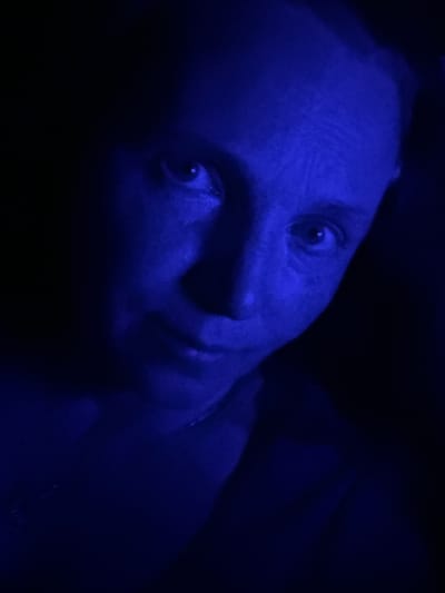 An individual's face is illuminated by a deep blue light in the foreground of a dark, indistinct environment. The subject's right eye is clearly visible, looking slightly off-camera. Their skin has a slightly textured appearance due to the lighting. The background is almost entirely black, with the blue light creating subtle gradients on the subject's features and the surrounding area. No specific location within McMasterville, Canada, can be identified from the image.