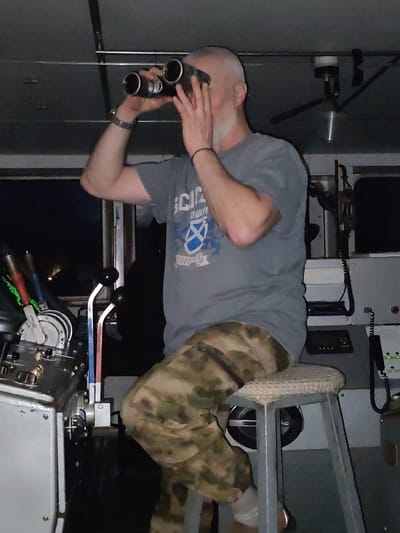 A man is sitting on a stool inside a cabin and looking through binoculars. The man has a shaved head, a gray T-shirt, and camouflage pants. The stool has a padded seat and metal legs. There are various controls and devices visible in the cabin, including levers, gauges, and a radio. The location appears to be a boat or ship, based on the equipment and the darkness outside the window. There is no information in the image to suggest it is in Tikhonovo, Russia.
