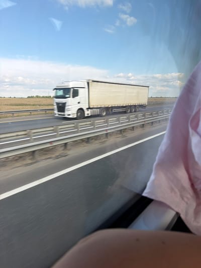 This image depicts a white semi-trailer truck driving on a highway. The truck is in motion, indicated by motion blur. The truck is white with a light-colored trailer. It is traveling on a multi-lane highway with a guardrail separating the lanes. The highway appears to be in a rural or semi-rural setting, with fields visible in the background. The sky is blue with scattered clouds. A portion of a pink garment and a person's arm are visible in the foreground, suggesting the photo was taken from inside a vehicle. The location is stated to be Pyatnitsa, Russia, though this cannot be independently verified from the image alone.